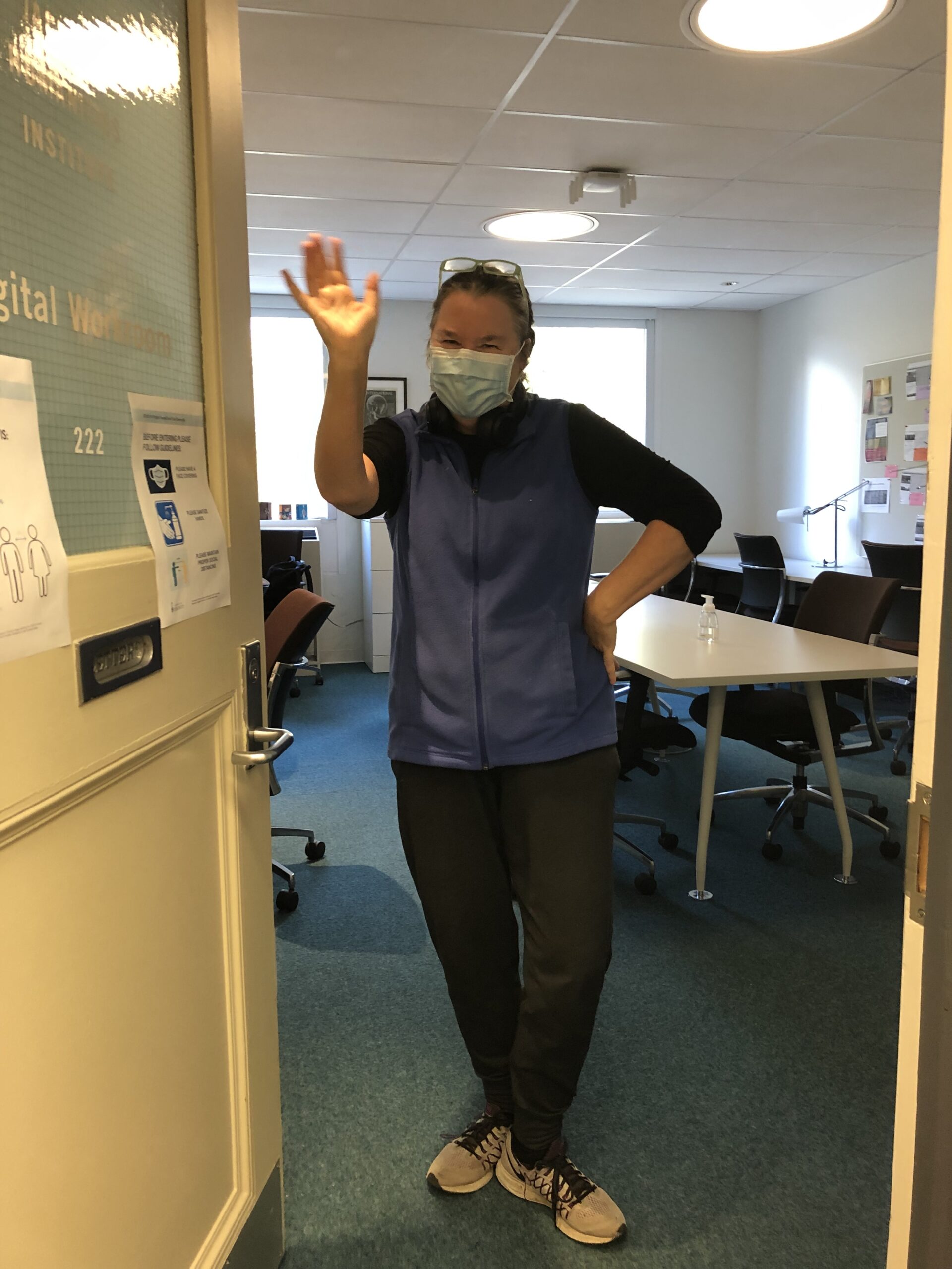 Elspeth Brown wearing a mask stands in the doorway and waves