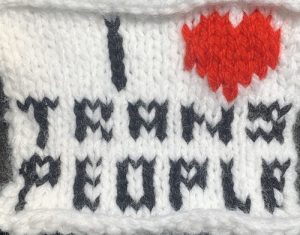  White knit panel with black text that says “I,” followed by a small red heart, “TRANS PEOPLE.”