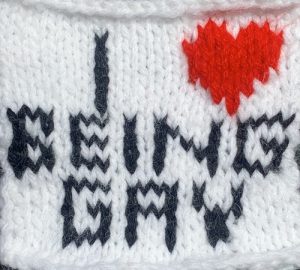 White knit panel with black text that says “I,” followed by a small red heart, “BEING GAY.”