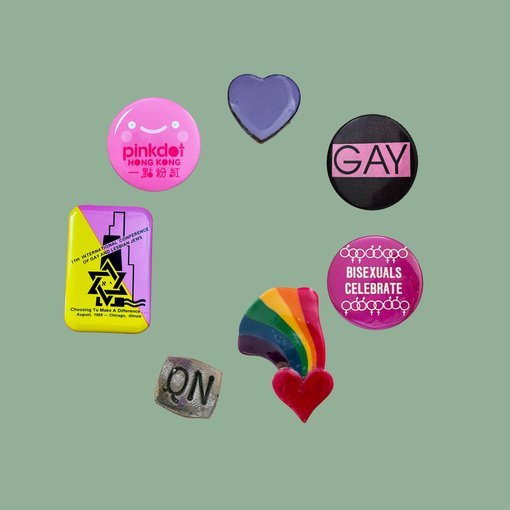 seven buttons. clockwise starting from the top:

1) heart-shaped, lavender (CB2040)

2) circular, black, pink rectangle in the centre has black text: “GAY” (CB2005)

3) circular, magenta with white text “BISEXUALS CELEBRATE”, two rows of white alternating male and female symbols respectively above and below text (CB2235)

4) red heart with rainbow (purple, blue, green, yellow, orange, red) flowing up from heart (CB1740)

5) rectangular, grey, “QN” carved in centre (CB2096)

6) rectangular with rounded edges, pink and yellow diagonal halves, black text: “11TH INTERNATIONAL CONFERENCE OF GAY AND LESBIAN JEWS” “Choosing To Make A Difference…” “August, 1989 — Chicago, Illinois”, black stylised abstract shapes and Star of David (CB1746)

7) circular, light pink, has a smiley face with a blush, magenta text: “pinkdot HONG KONG 一點粉紅“ (CB1790)